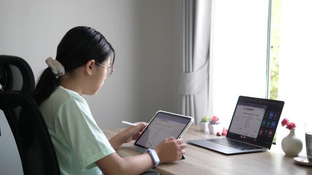 Cinematic wide over-the-shoulder shot of a young female creative professional using a stylus on digital tablet, multitasking with laptop on desk