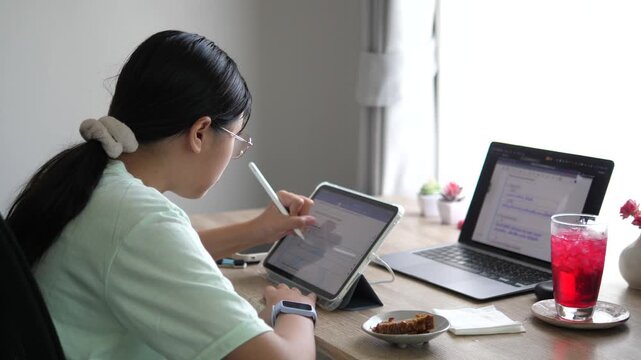 Cinematic wide over-the-shoulder shot of a young female creative professional using a stylus on digital tablet, multitasking with laptop on desk