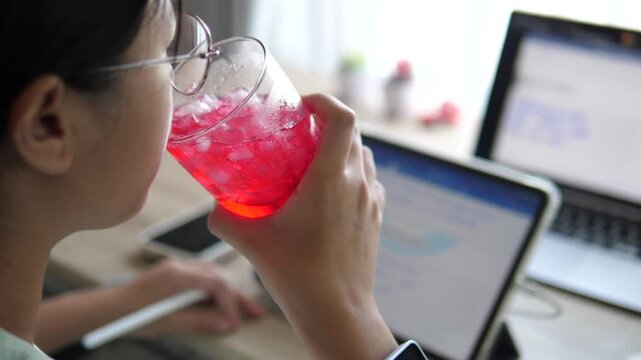 Cinematic over the shoulder shot of a young female professional with glasses drinking a refreshing red iced beverage while working with laptop and tablet on desk