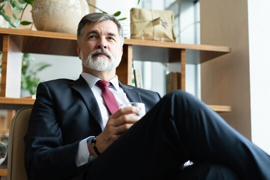 Mature bearded businessman in formal wear drinking fresh water while sitting in the modern office