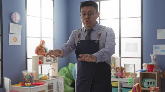Man teacher in apron finger to lips for silence and extends hands over toys in kindergarten building; calm patience.