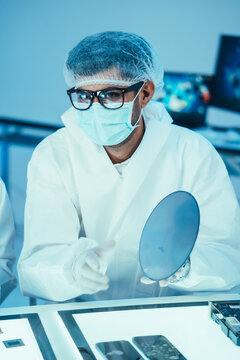 Male engineer in a protective cleanroom suit and face mask holding and inspecting a silicon wafer disc for semiconductor microchip manufacturing in a high tech laboratory setup