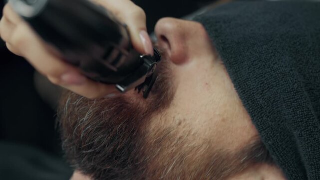 Close up of barber trimming mustache with electric clipper during beard grooming procedure in modern barbershop