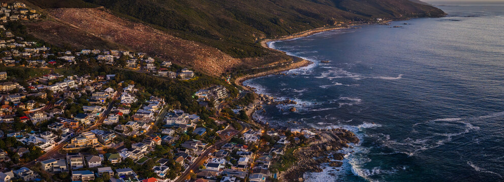 Aerial view of Cape Town at sunset shows Camps Bay and Clifton, winding cliffside roads, terraced homes on slopes, waves on rocky shore, and the Twelve Apostles range.