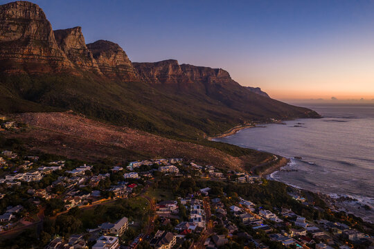 Aerial view of Cape Town at sunset shows Twelve Apostles and Camps Bay. Westward horizon glows purple orange as surf meets rocky coast and hillside homes nestle.