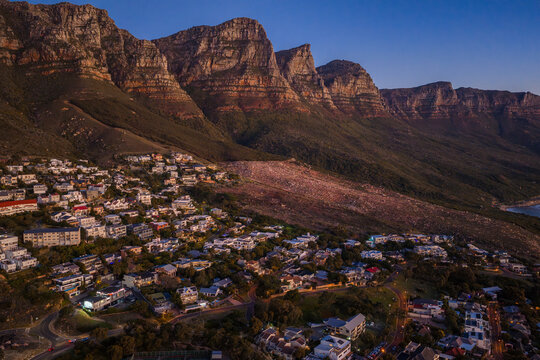 Aerial sunset view of Camps Bay and Clifton in Cape Town, South Africa, with terraced neighborhoods climbing to the Twelve Apostles as city lights start to twinkle.