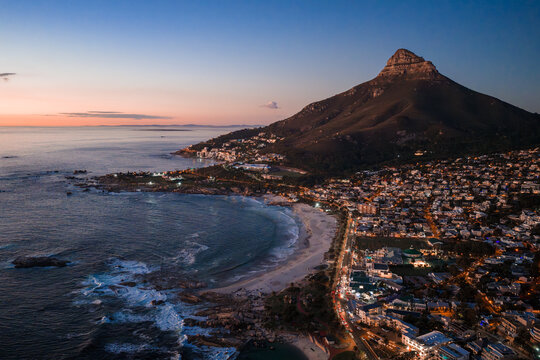 Aerial view of Cape Town, South Africa, at dusk shows Lion's Head, Camps Bay, Clifton beaches, waves on rocks, coastal road, packed homes, and emerging city lights.