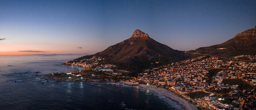 Aerial view of Cape Town, South Africa at dusk shows Lion's Head, the Atlantic, Camps Bay, and Clifton as lights glow along the curving shore under fading purple and rose tones.