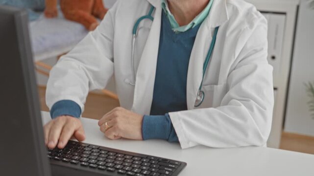 Man doctor with stethoscope wearing glasses, typing at keyboard beside stuffed toy in clinic building; thoughtful care.