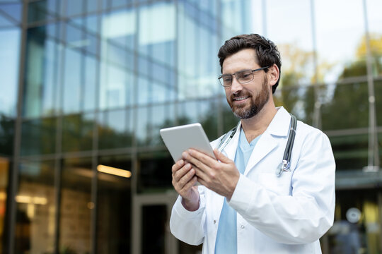 Smiling male doctor wearing a white coat and stethoscope using a digital tablet for healthcare technology outdoors in front of a modern clinic, representing telehealth and medical innovation