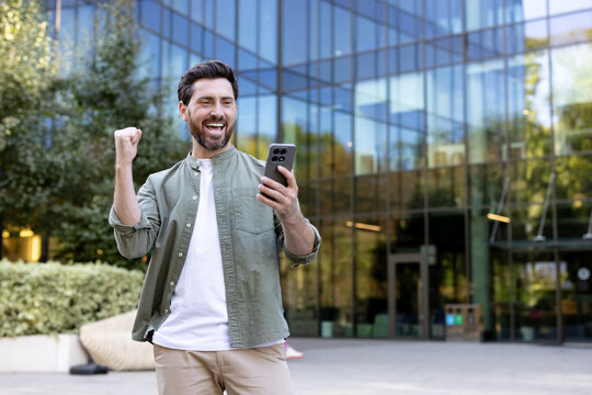 Happy man looking at his smartphone, raising a fist in triumph, receiving excellent news or achieving success in front of a modern glass office building outdoors
