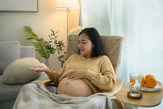 Asian Pregnant Woman Taking Prenatal Vitamins at Home While Relaxing in Comfortable Chair
