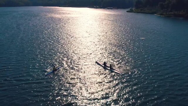 Drone shot of rowers moving across sunlit lake water with bright reflections and ripples. Peaceful aerial scene showing outdoor sport, endurance, teamwork, freedom and nature.