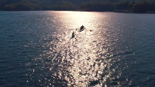 Drone shot of kayakers and rowers moving across sunlit lake water with bright reflections and ripples. Scenic outdoor sport scene showing teamwork, training, freedom and nature.
