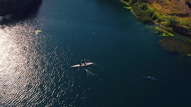 Drone shot of a kayaker and rowing team moving across a sunlit lake near a natural shoreline. Scenic aerial view with ripples, reflections, freshwater, outdoor sport and nature.