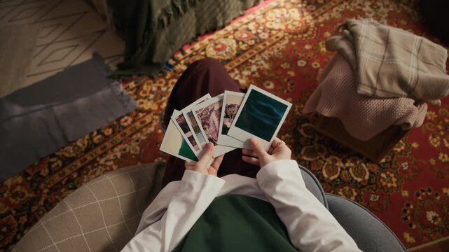 Top view of teenage girl sitting in armchair and holding instant polaroid photographs in hands, creating nostalgic atmosphere in warm cozy bedroom interior