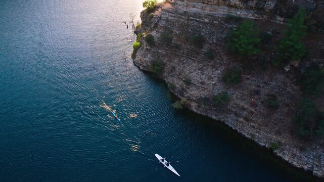 Drone shot of a kayaker and rowing boat moving beside a rocky lake shoreline with clear blue freshwater. Scenic outdoor sports scene with cliffs, ripples, nature and calm motion.