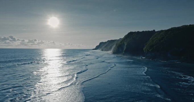 Indian Ocean waves breaking along the green cliffs of Bali's Bukit Peninsula at evening, bright sun reflecting on water surface creating serene tropical travel destination landscape. Aerial drone shot