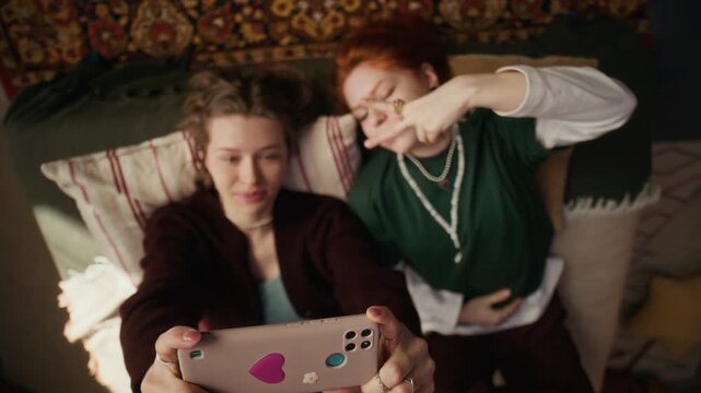 Overhead view of two young Gen Z female friends lying on pillows on bed while taking funny selfie with smartphone in cozy bedroom