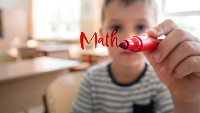 Young child writing math word with red marker in focus, blurred classroom background and playful learning environment, educational schoolboy creative classroom concept