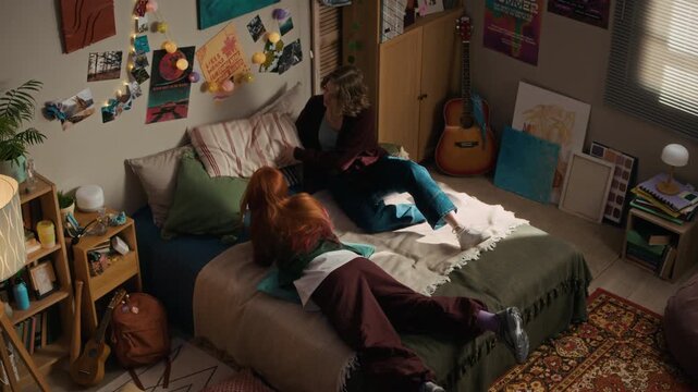 Overhead perspective of young female friends spending time together lying down on bed and browsing mobile phone in cozy bedroom apartment