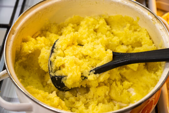 High angle view of hot freshly boiled yellow potatoes mashed inside traditional enameled kitchen pot using black plastic hand masher tool during home cooking process.