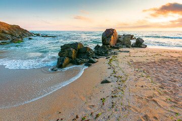 Fototapeta premium huge stones on the sandy beach at sunrise. wonderful holiday season background at the sea. clouds on the blue sky above horizon. view of waves washing the shore. scenic seascape for relaxation