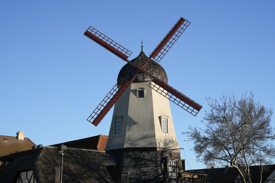Traditional windmill with red blades and white tower rising above village rooftops under a clear blue sky, evoking European architecture, heritage tourism, and historic rural technology.