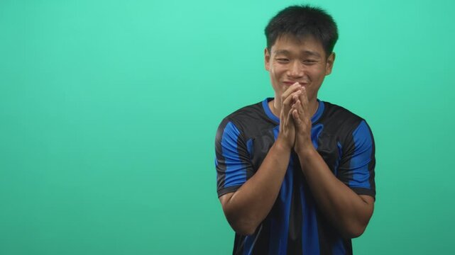 Man in blue and black soccer uniform clasps hands near chest while smiling with a sheepish shrug in studio; amused humility.