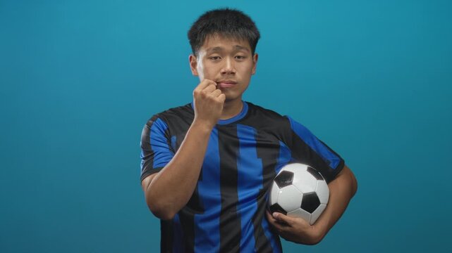 Young man in a blue and black soccer uniform holds a ball under his arm and places finger to lips in studio; thoughtful quiet contemplation.