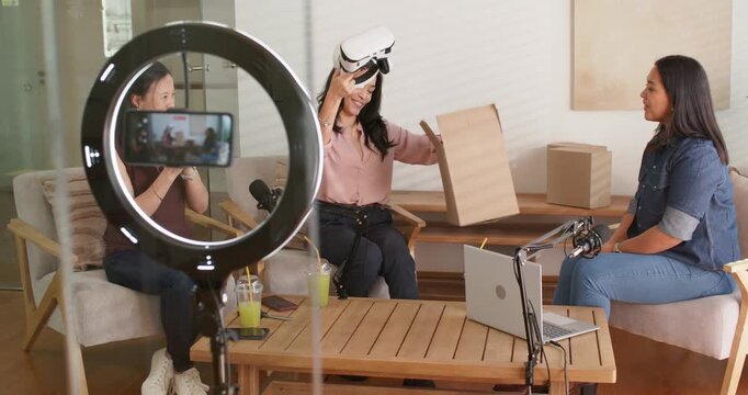 Diverse female hosts in studio, center Asian woman getting box opening showing VR headset to camera