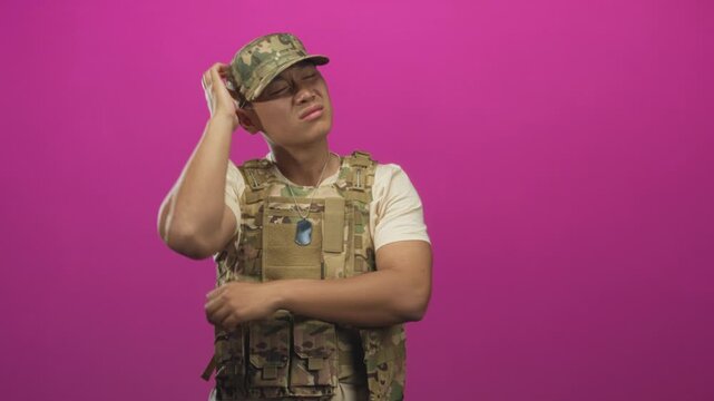 Man in camouflage uniform and tactical vest touches temple with hand while holding dogtag in studio; contemplation duty.