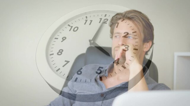 Mid-adult man sitting at desk clock hands advancing rubbing chin reviewing laptop for business task