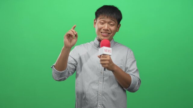Young man holding a news microphone with red windscreen, right hand gripping mic and speaking to camera in studio; confident reporting.
