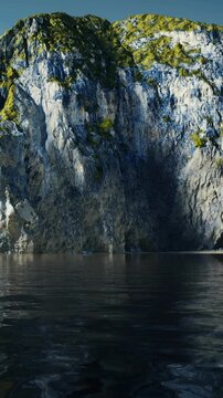Rock cliffs rise above calm water, showing textures and colors. The scene captures natural features and clear reflections. This view is taken in the evening light near the waters edge.