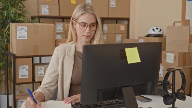 Woman writing with pen at computer among stacked parcels, boxes and shipping labels in a small fulfillment building; focused productivity.
