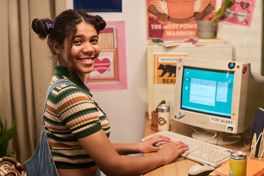 Portrait of teenage girl using desktop computer at home desk, smiling at camera while typing, representing digital learning and youth technology. Useful for education or teen tech campaigns