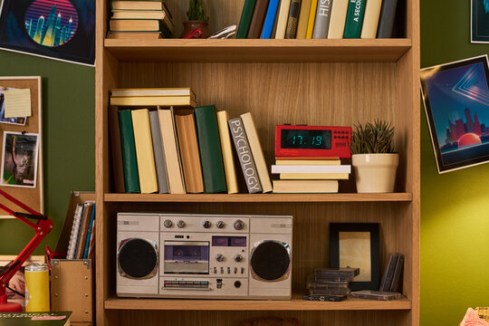 Bookshelf displaying books, retro boombox, cassette tapes, digital clock, plant, framing nostalgic home media and study culture. Useful for education, music, lifestyle, vintage marketing