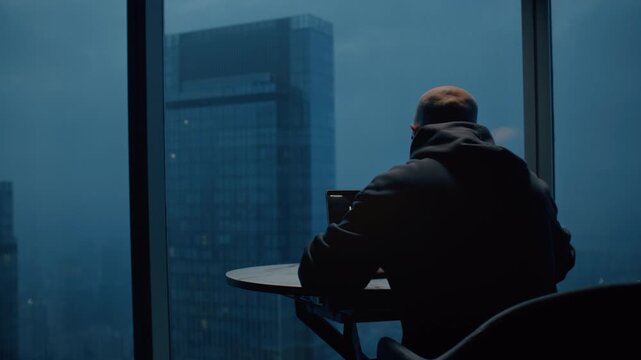 Bald man trader trades on stock exchange on laptop at home at night, lives in a skyscraper, a night city. Male investor analyzes stock market and cryptocurrencies. Apartment in downtown. BTC Ethereum
