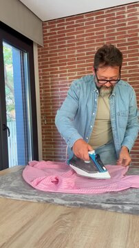 A man meticulously irons his pink shirt at home, completing a common domestic chore with care and focus, showcasing an everyday lifestyle scene.