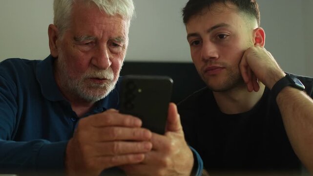 An older man and a younger man share a moment, looking at a smartphone together, connecting through technology and intergenerational learning. They appear focused and engaged.