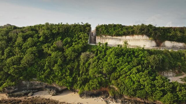 Aerial drone flight into limestone cliff path, rock canyon road on Bukit Peninsula tropical remote beach, surrounded by lush green forest, and shallow coral reef, creating a dramatic landscape in Bali