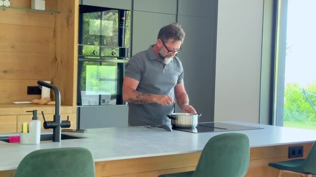 Mature man preparing a meal in a modern kitchen. He places a pot on the induction cooktop and smiles, enjoying his time at home.