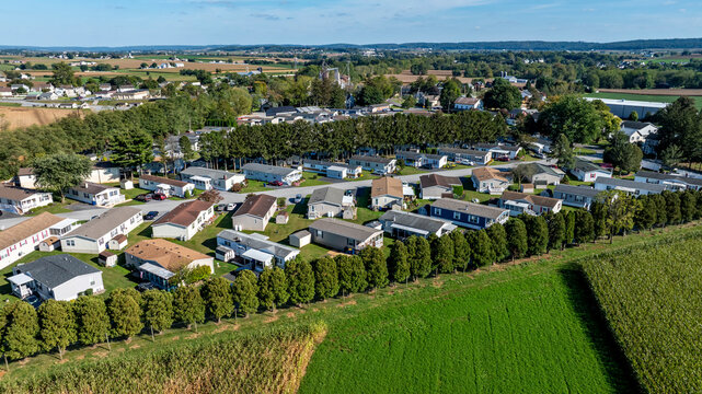 A collection of mobile, manufactured homes aligned in rows in a rural community. The houses are arranged in a neat pattern with trees and open fields nearby. Farms are visible in the distance.