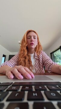 A young woman with red hair experiences frustration while working on her laptop, rubbing her temples in an attempt to alleviate stress from a challenging task or problem.