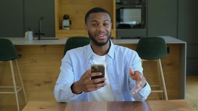 A happy young Black man holds his smartphone while speaking and gesturing to the camera, engaging in a video call from a modern home kitchen.