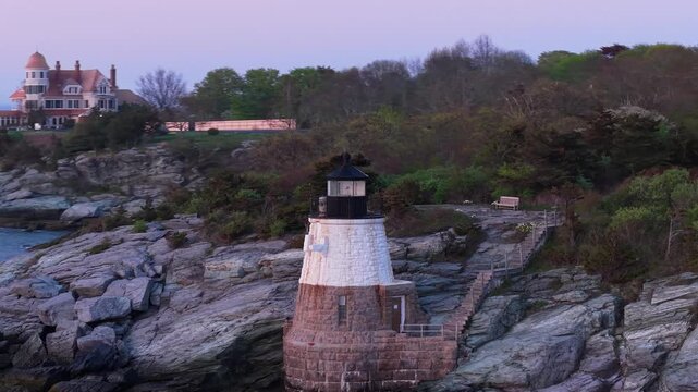 Castle Hill Lighthouse at sunset aerial video, a scenic drone shot of the historic beacon illuminated on a rocky cliff point during a vibrant twilight in Newport, Rhode Island.