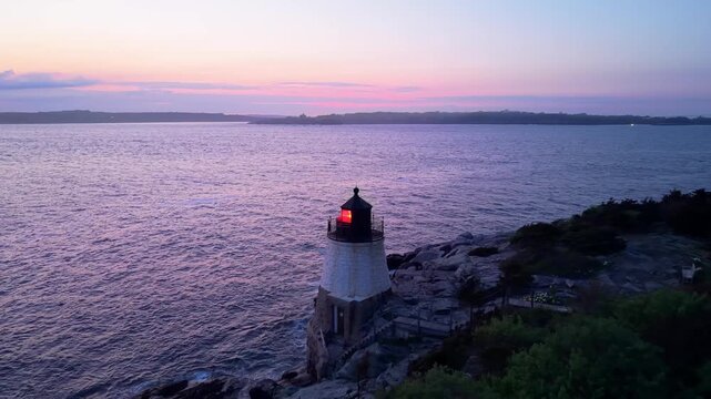 Castle Hill Lighthouse at sunset aerial video, a scenic drone shot of the historic beacon illuminated on a rocky cliff point during a vibrant twilight in Newport, Rhode Island.