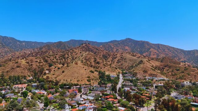 Sunlit residential area with beautiful villas and green trees. Approaching the rocks with scarce vegetation. Los Angeles, California, USA.