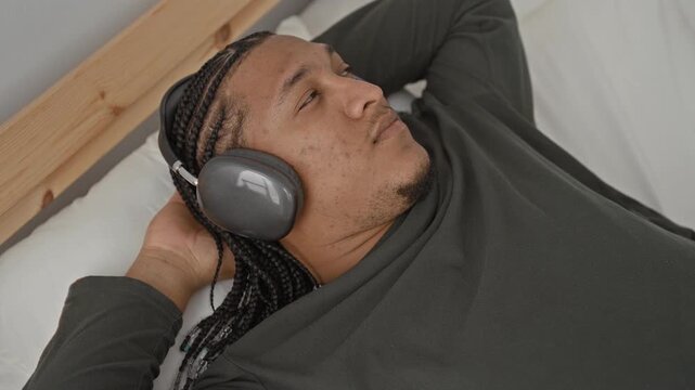Man wearing headphones lying on a bed with hand behind head and eyes closed in a bedroom, braided hair visible; relaxed introspection.
